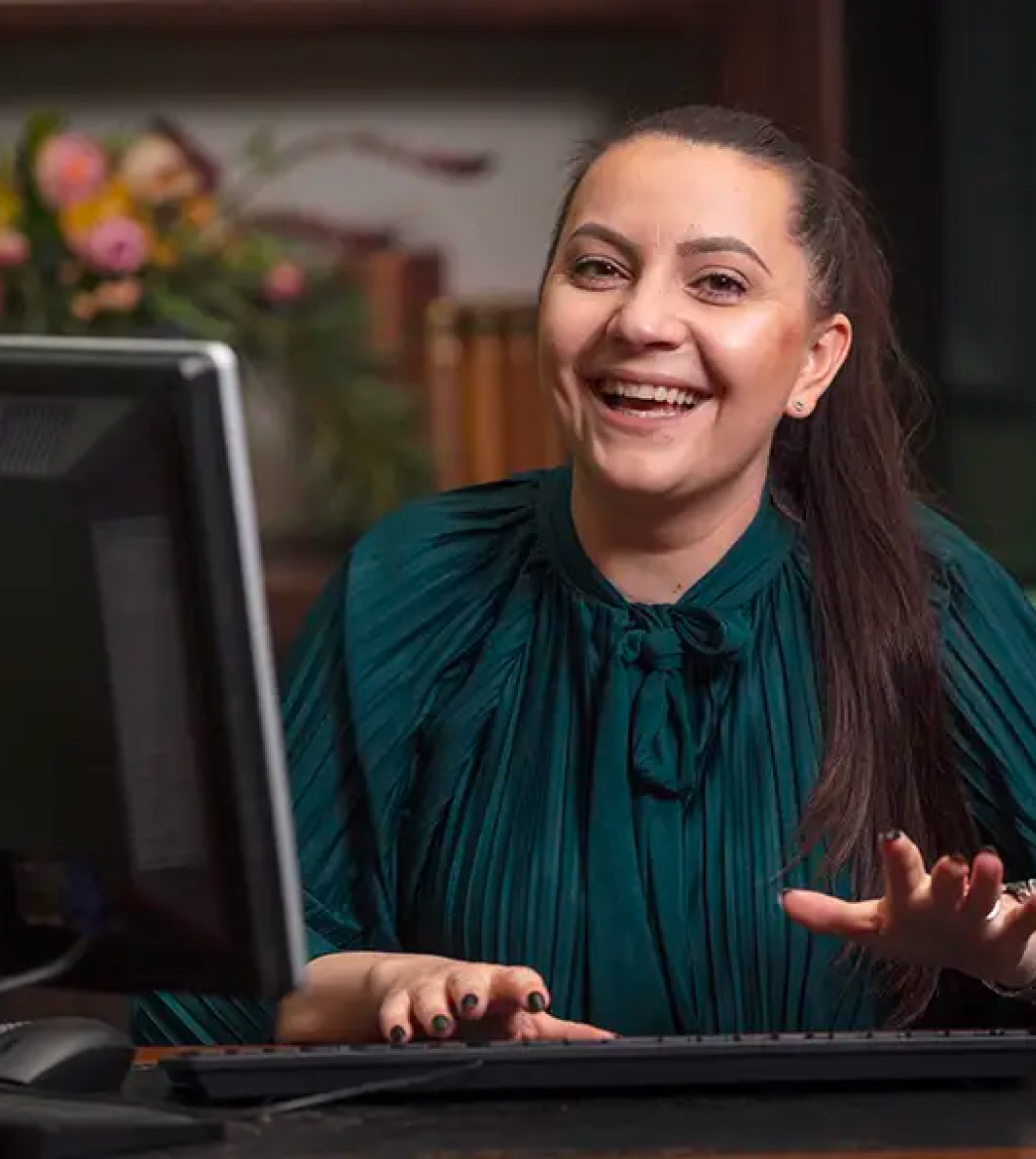 A woman in a green blouse sits at a desk, smiling, with hands on a keyboard. A computer monitor is visible. There are flowers and books in the background.