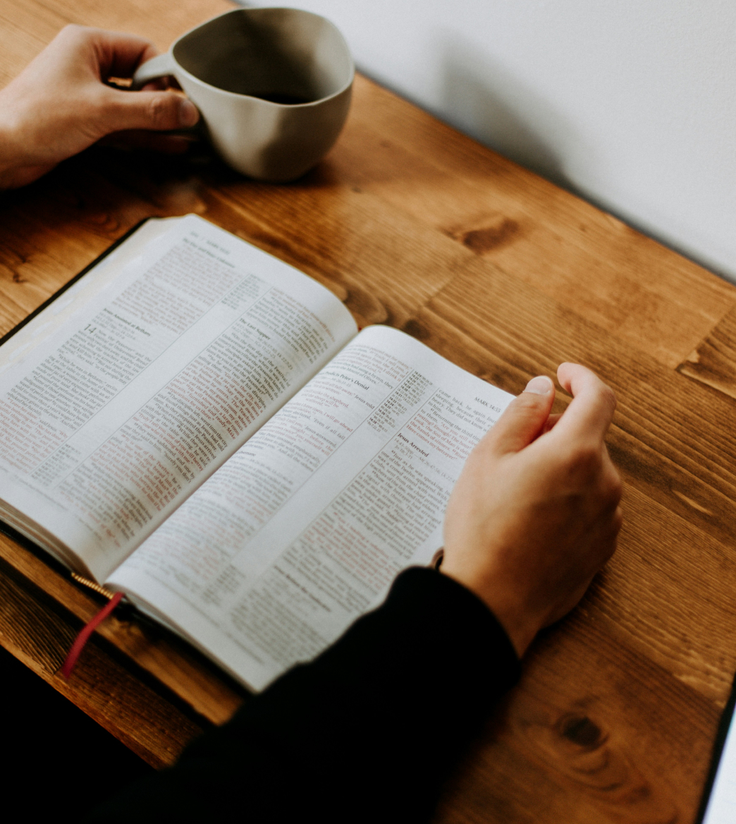 Person reading an open book on a wooden table while holding a cup.