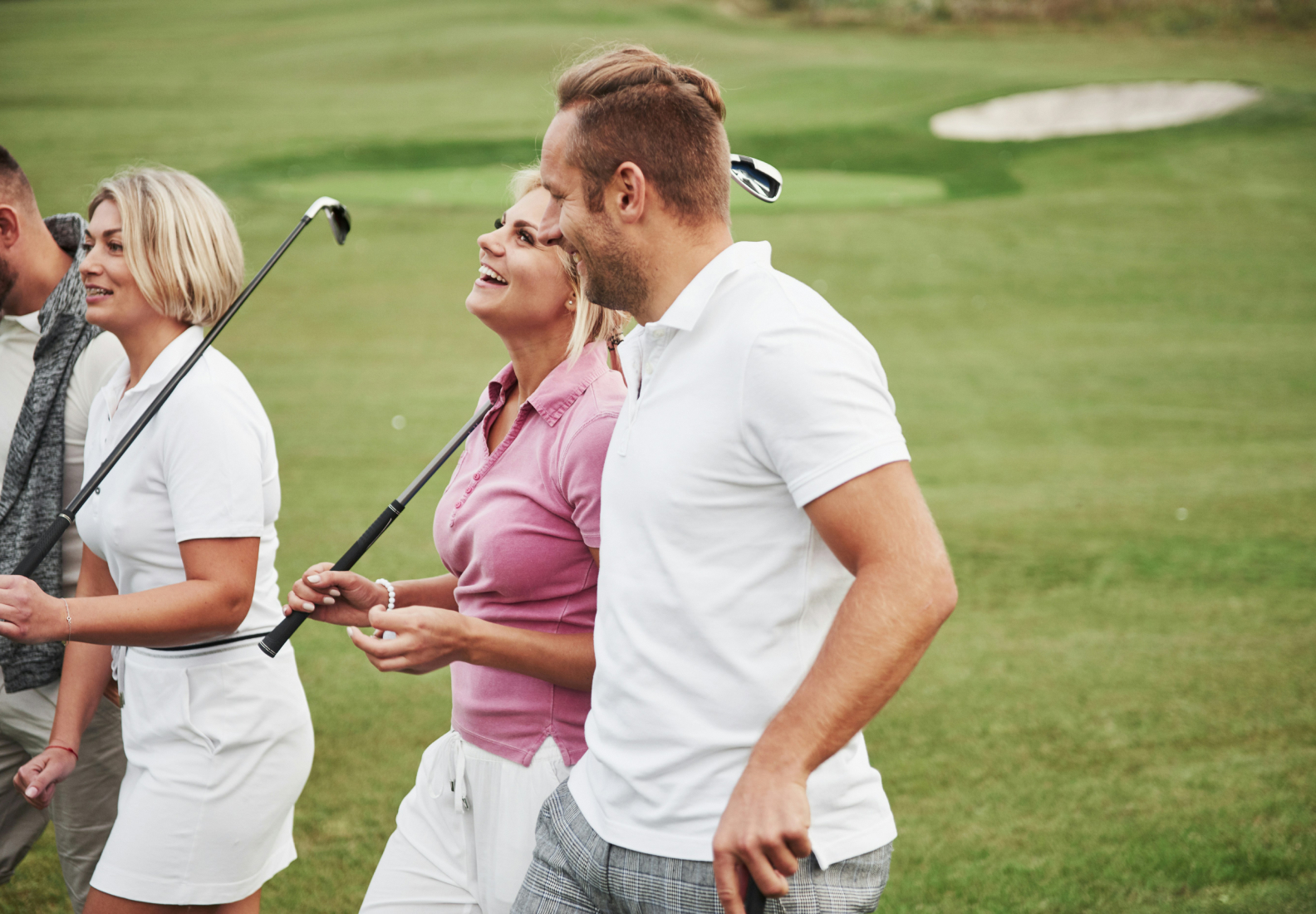 Four people walking on a golf course, holding clubs, and smiling.