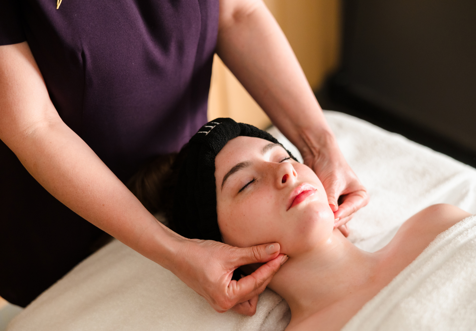 A person receives a facial massage while lying on a white towel. Hands are positioned on the person's jawline.