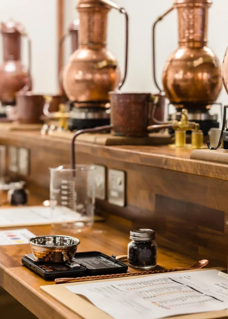 A distillery workspace with copper stills, a beaker, a jar, and equipment on a wooden surface.