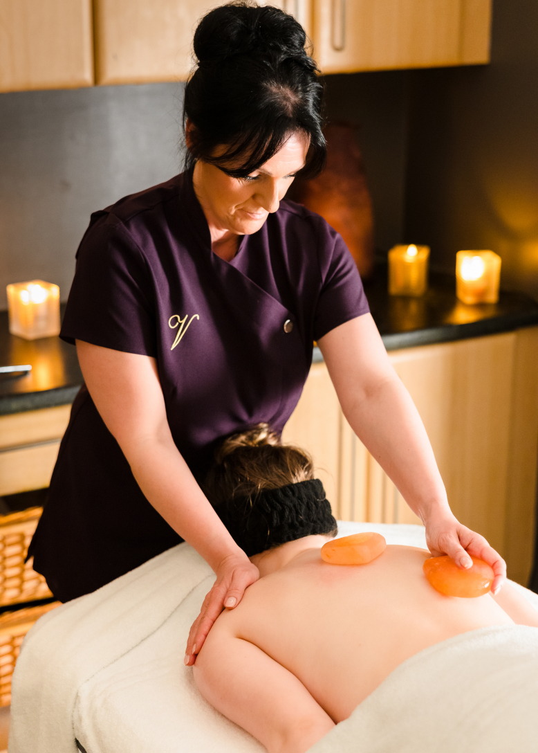 A massage therapist places warm stones on a client's back in a dimly lit spa room with candles.