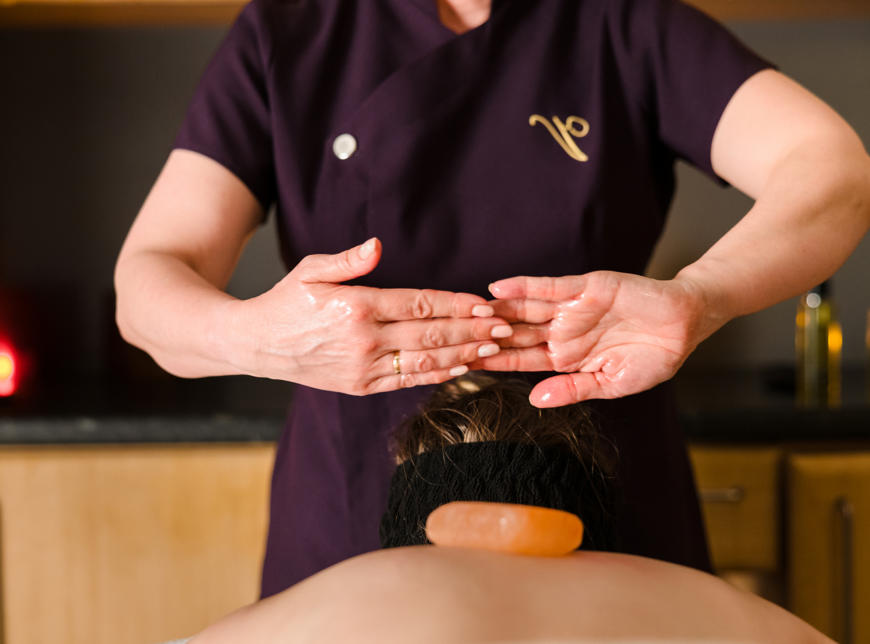 A person receiving a massage on their upper back with a stone placed on their spine. The massage therapist is wearing a dark uniform.