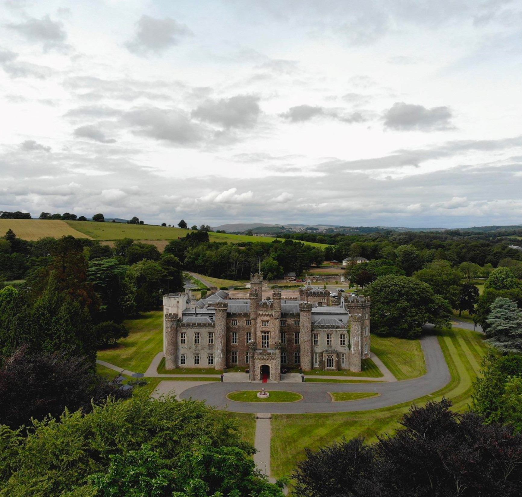 Aerial view of a large castle surrounded by lush greenery, with a circular driveway in the foreground and rolling hills in the background under a cloudy sky.