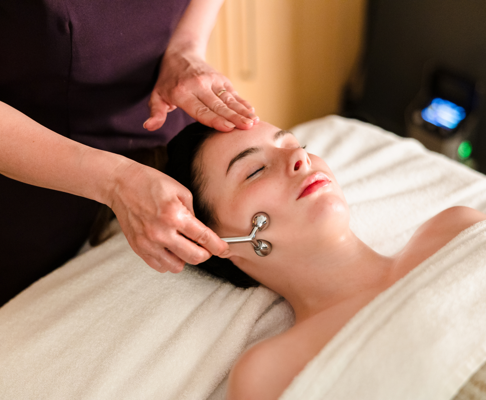 Person receiving a facial treatment with a metal roller device while lying on a padded table.