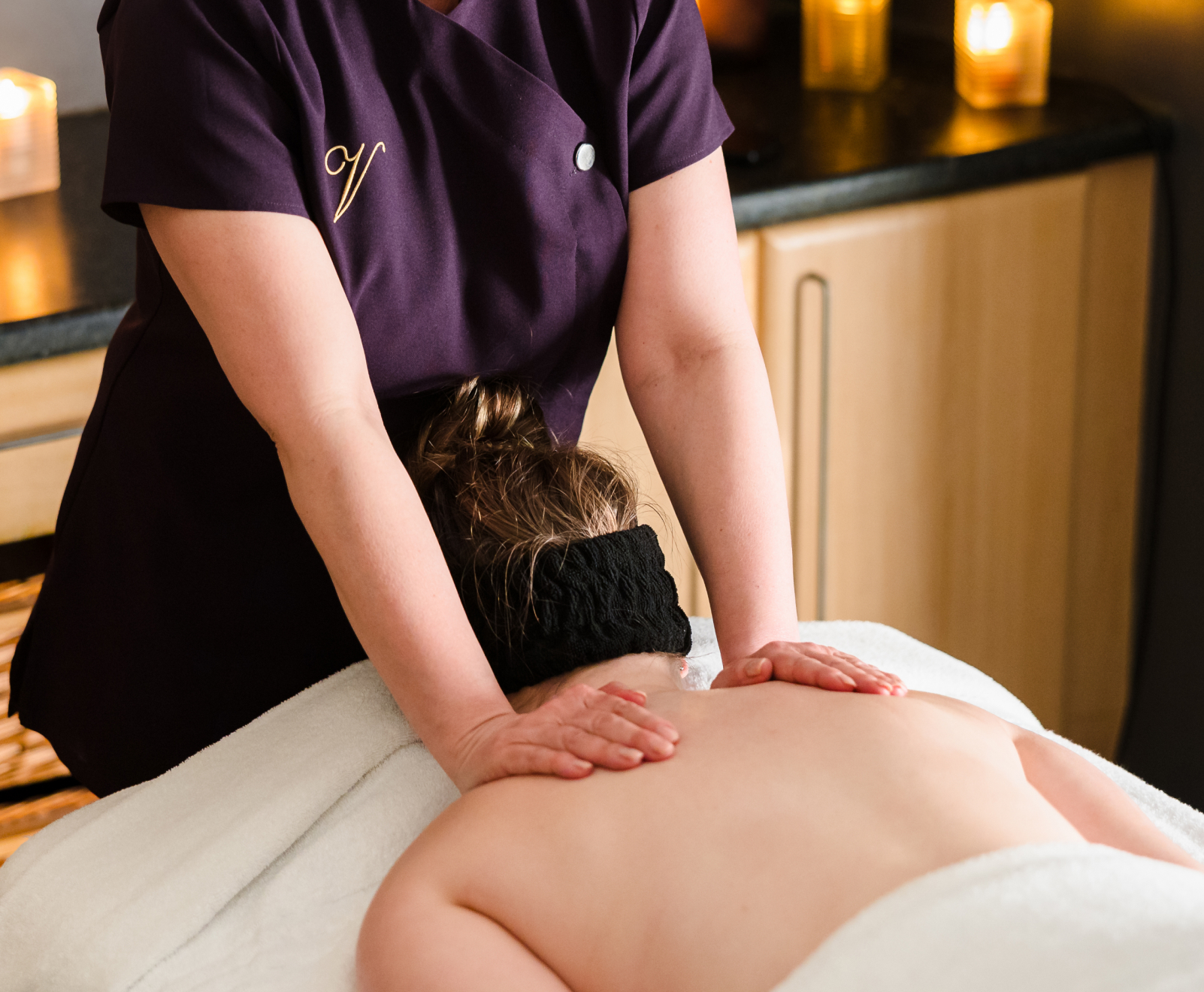 A massage therapist in a purple uniform gives a massage to a person lying face down on a table, with candles in the background.