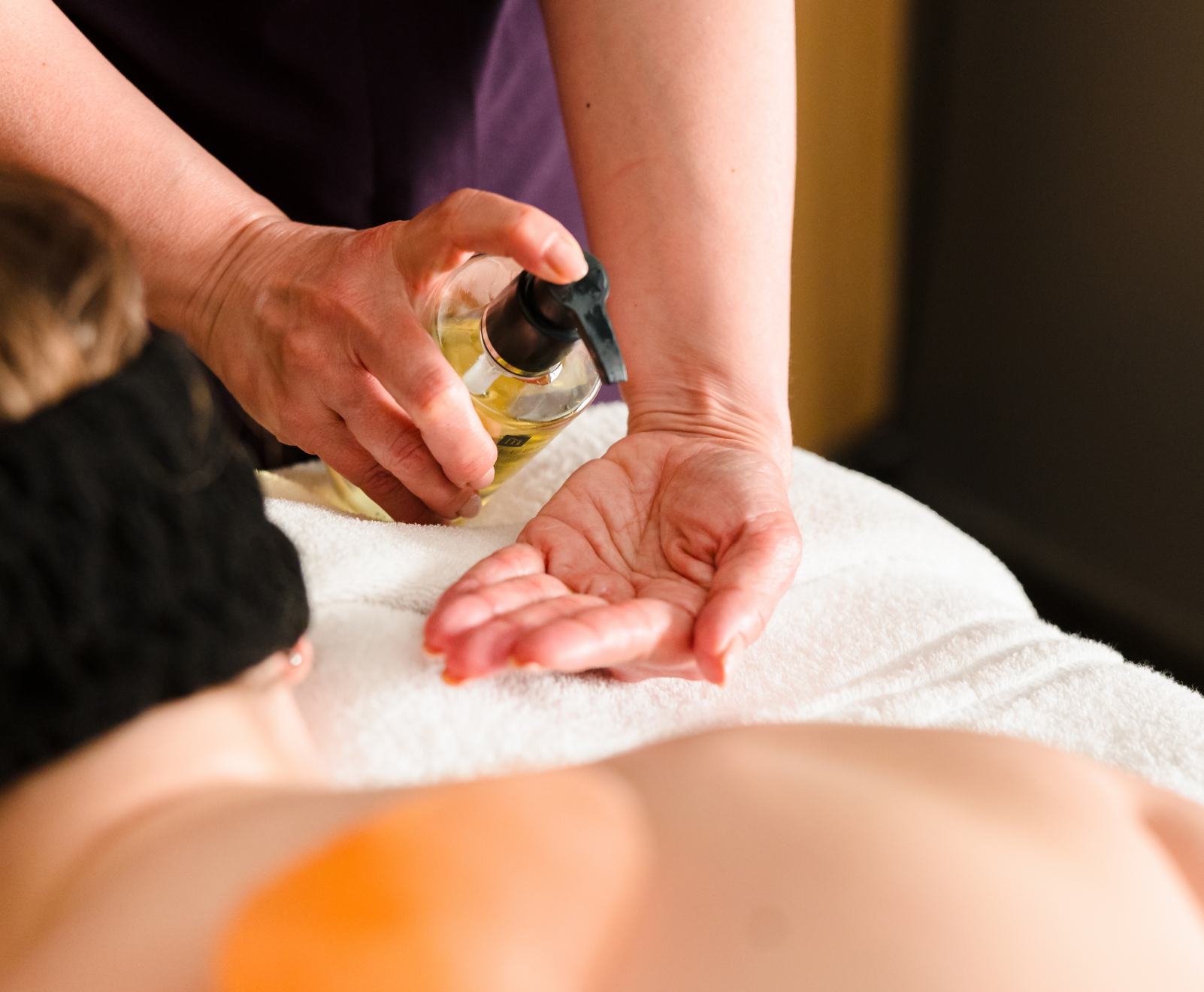 A person's hand pumps massage oil onto their palm over a towel-covered table, with another individual lying face down, blurred in the foreground.