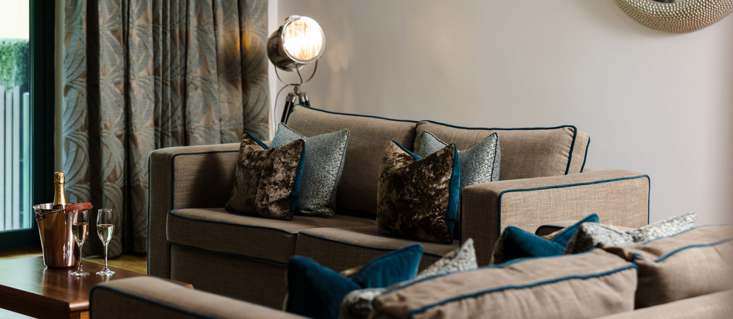 A modern living room with a beige sofa, patterned cushions, and a spotlight lamp. A champagne bottle and glasses are on a side table. Drapes cover a window, and a mirror is on the wall.