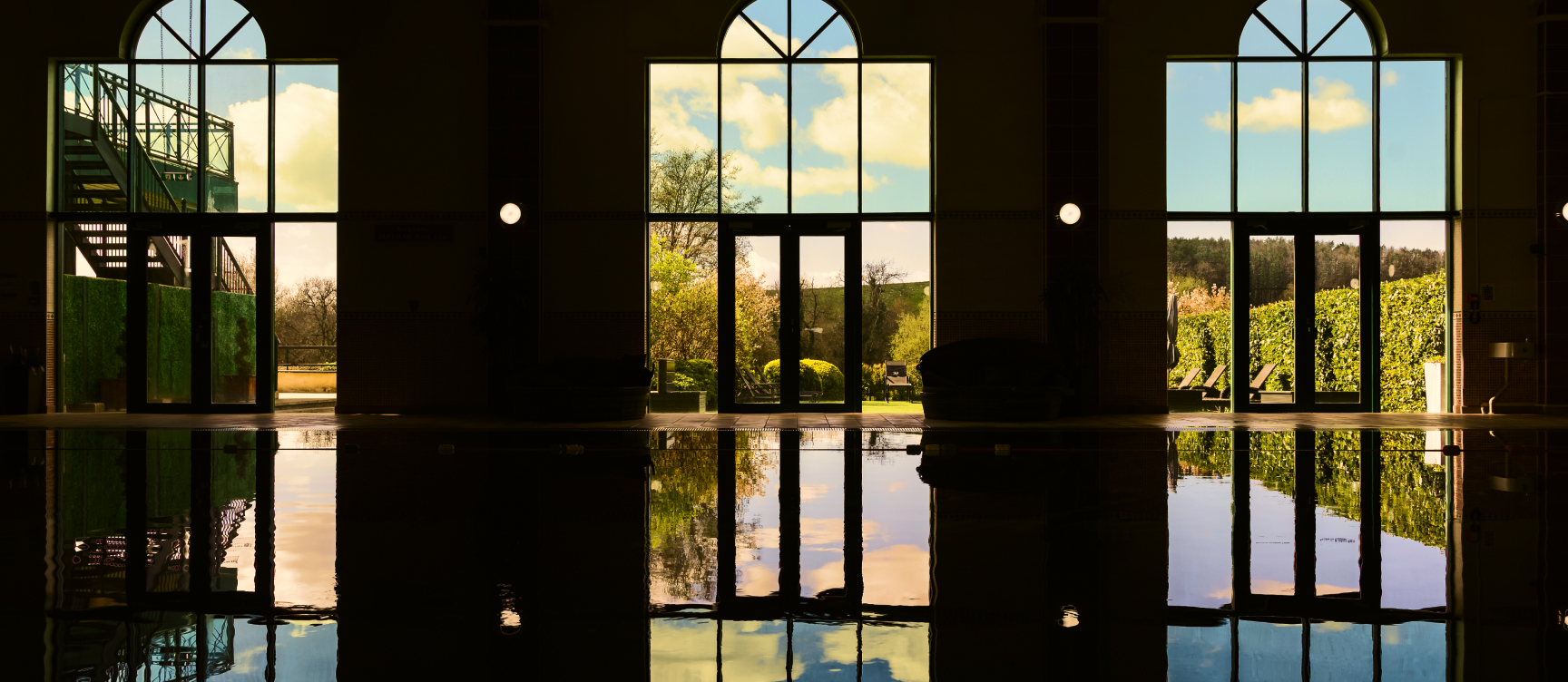 An indoor pool reflects light from large windows, revealing a garden outside. Arched doorways and a staircase on the left add architectural interest to the scene.