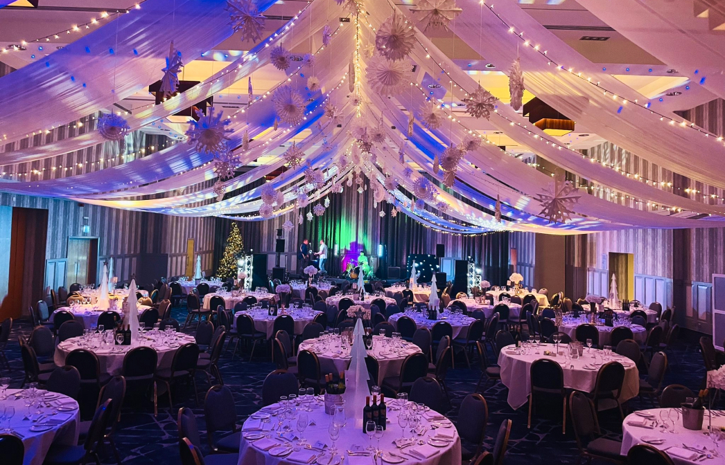 Ballroom with round tables set for an event, featuring draped ceiling decorations and colorful lighting.