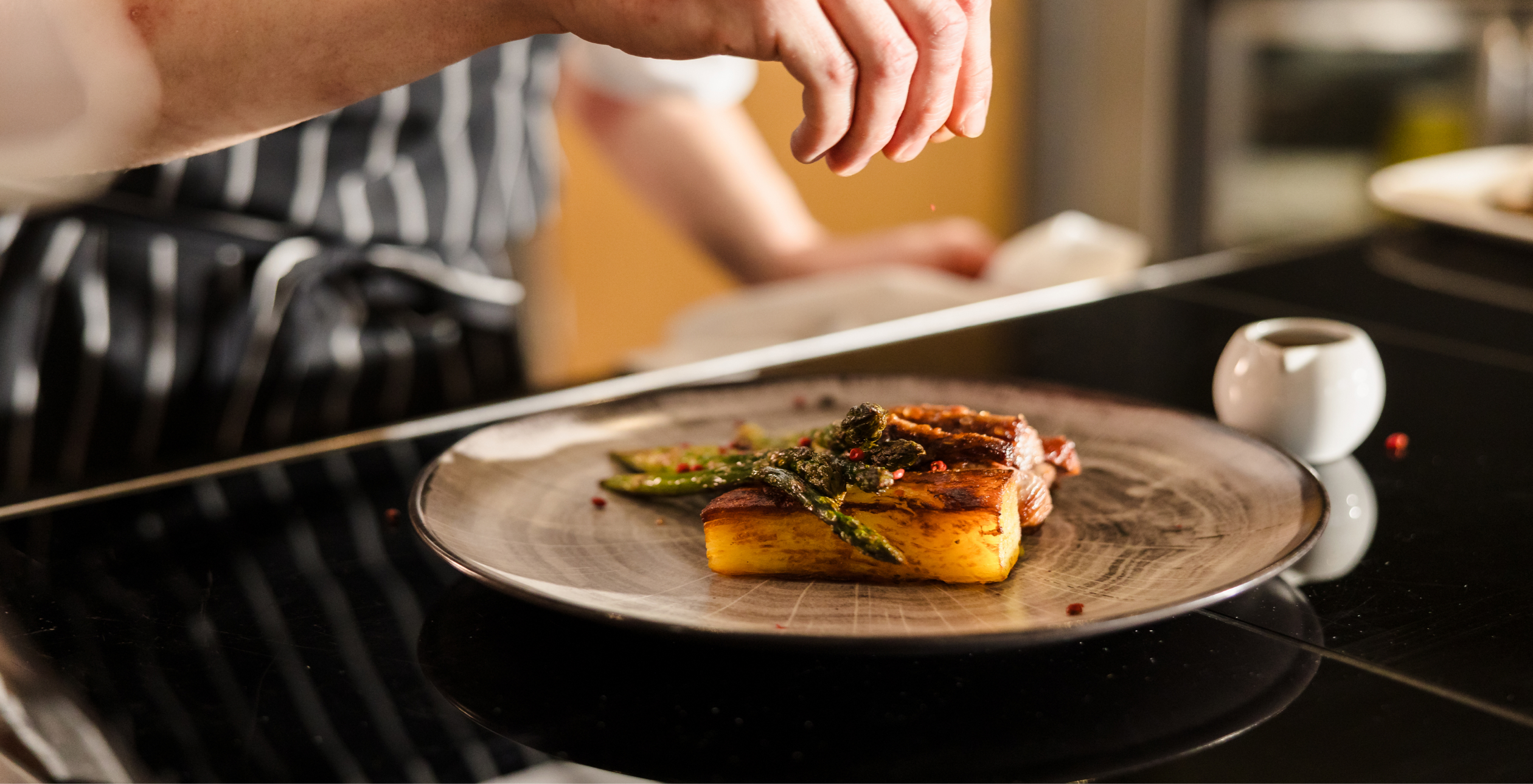 A chef is garnishing a plated dish with grilled polenta, vegetables, and sauce.