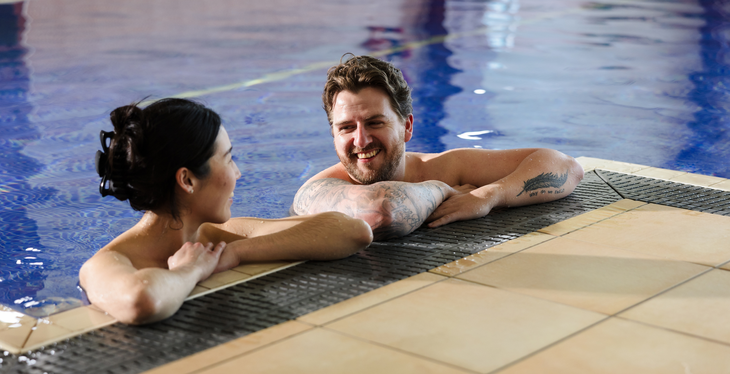 Two people are relaxing and talking at the edge of a swimming pool.