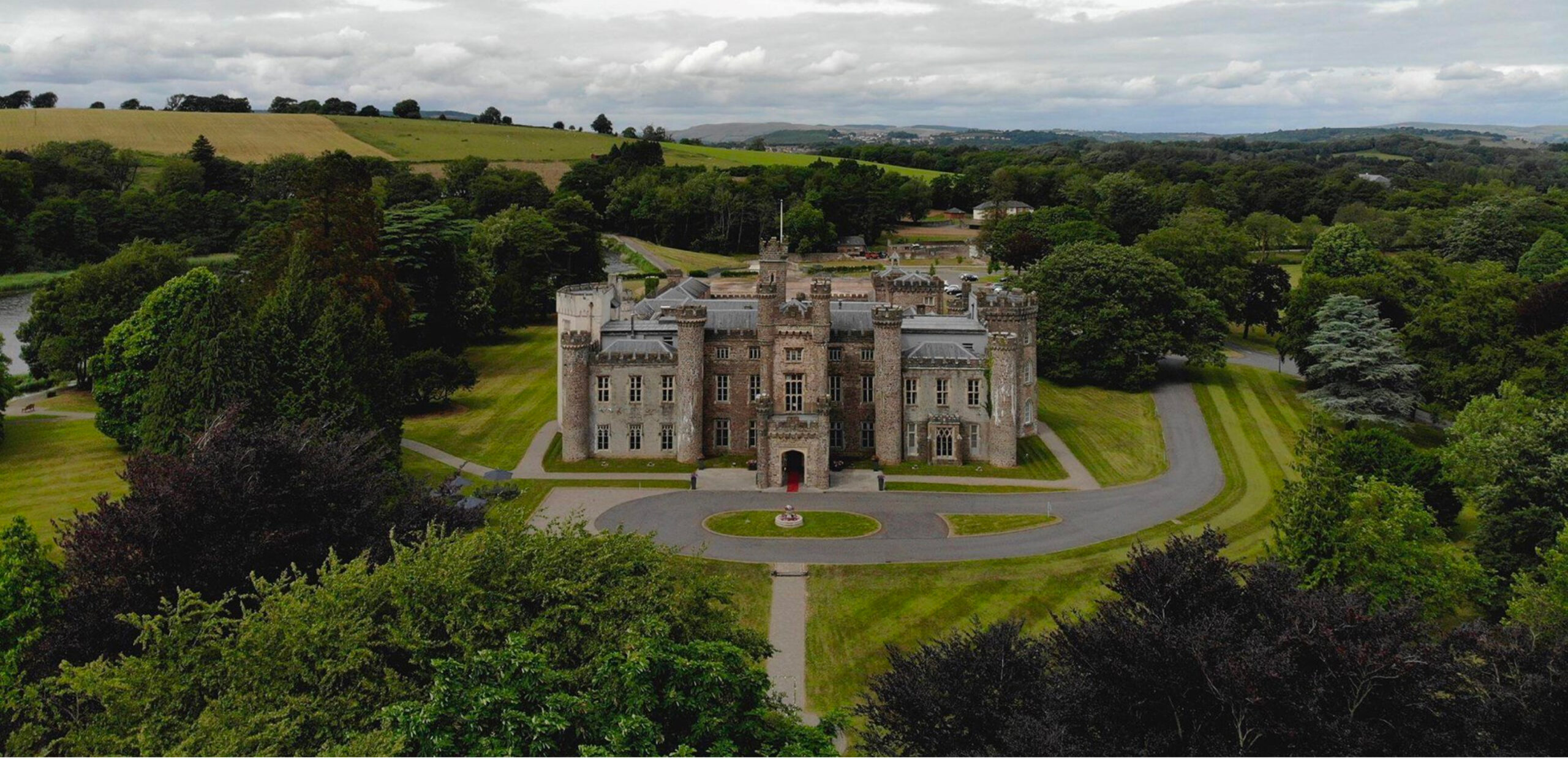 Aerial view of a historic castle surrounded by lush green trees and rolling hills, with a circular driveway in front.