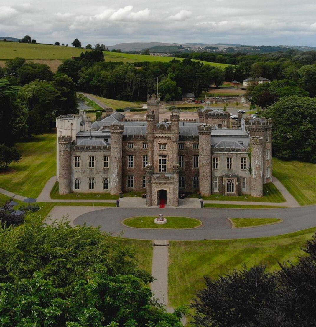 An aerial view of a historic stone castle surrounded by green fields and trees, with a circular driveway in front.