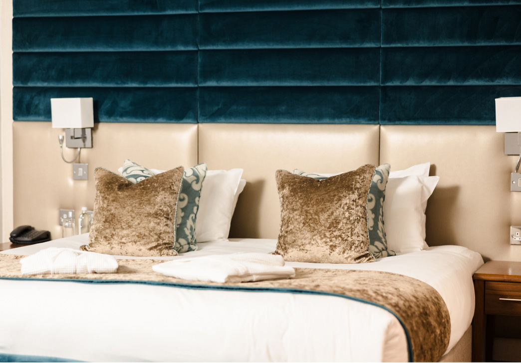 A hotel room with a large bed, accented by plush beige pillows and a blue padded headboard. Two wall-mounted lights are above the bed.