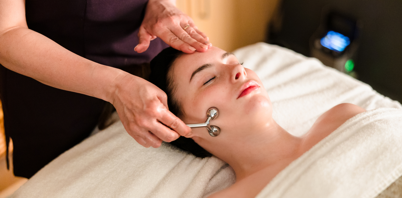 Person receiving a facial massage with a metal roller tool while lying on a massage bed.