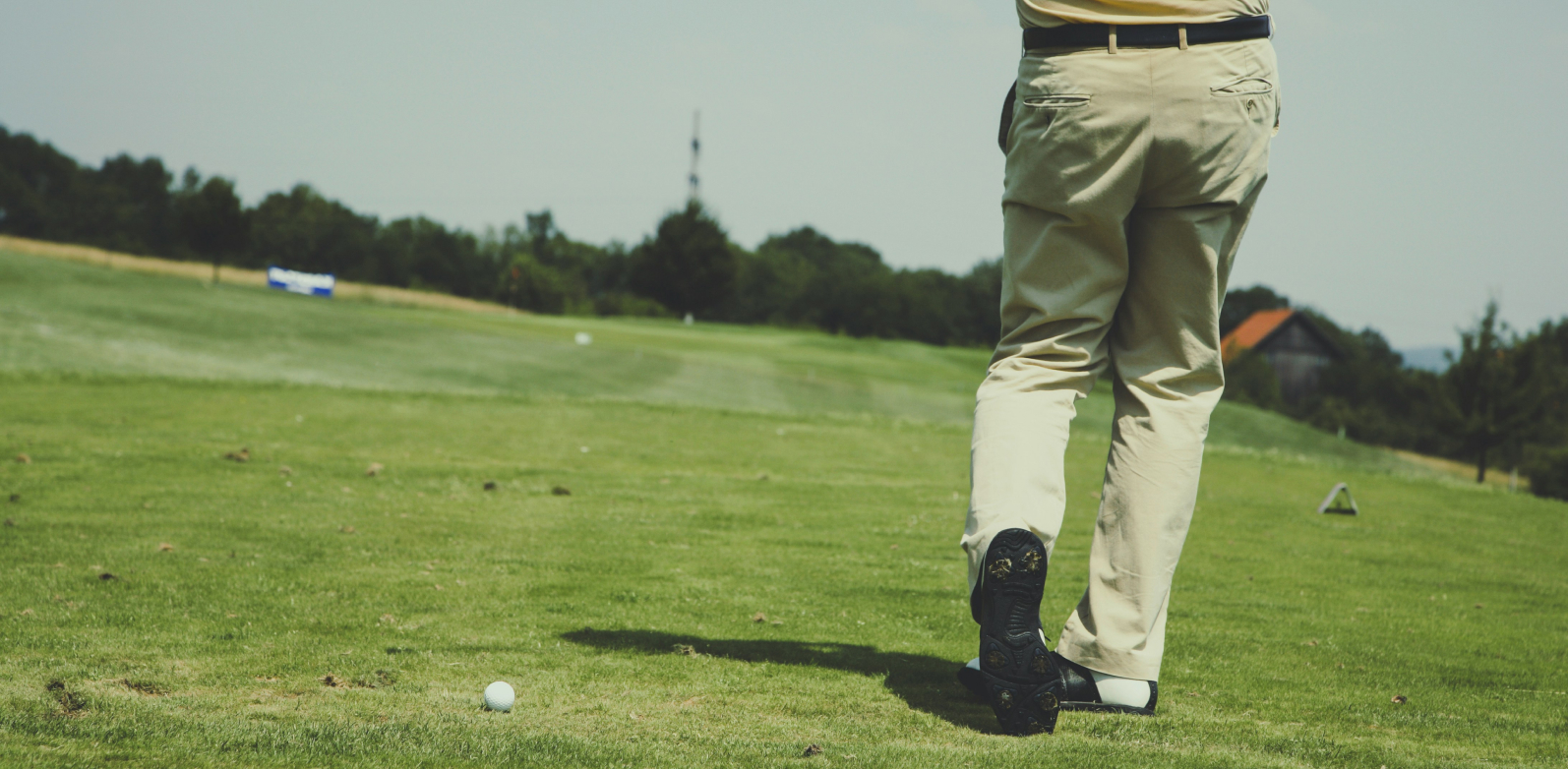 Person on a golf course, swinging a club towards a ball on the green. Distant trees and a small building are in the background.