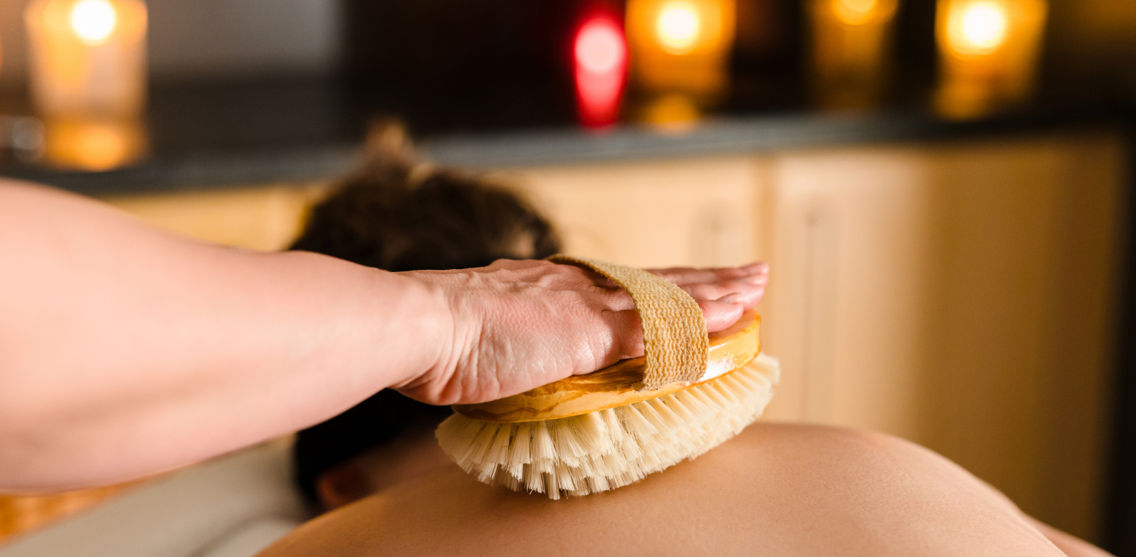 A person receiving a back massage with a brush in a dimly lit room with candles in the background.