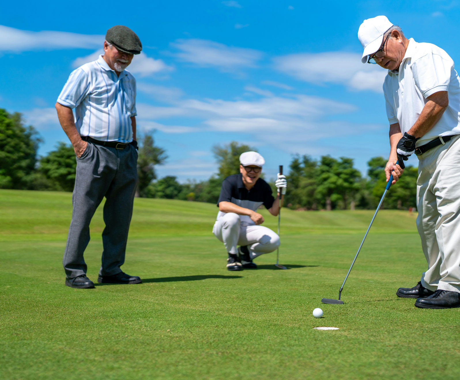 Three men on a golf course, one putting while the others watch. They're in casual golf attire, with blue sky and trees in the background.