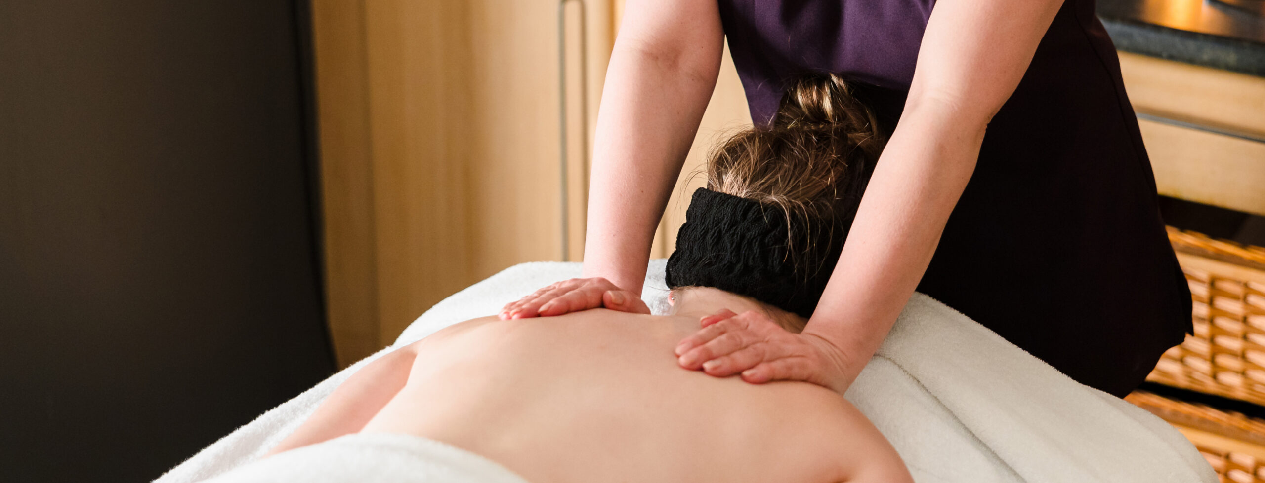 A person receives a back massage in a spa setting, lying on a table with a towel, while the masseuse applies pressure with both hands.