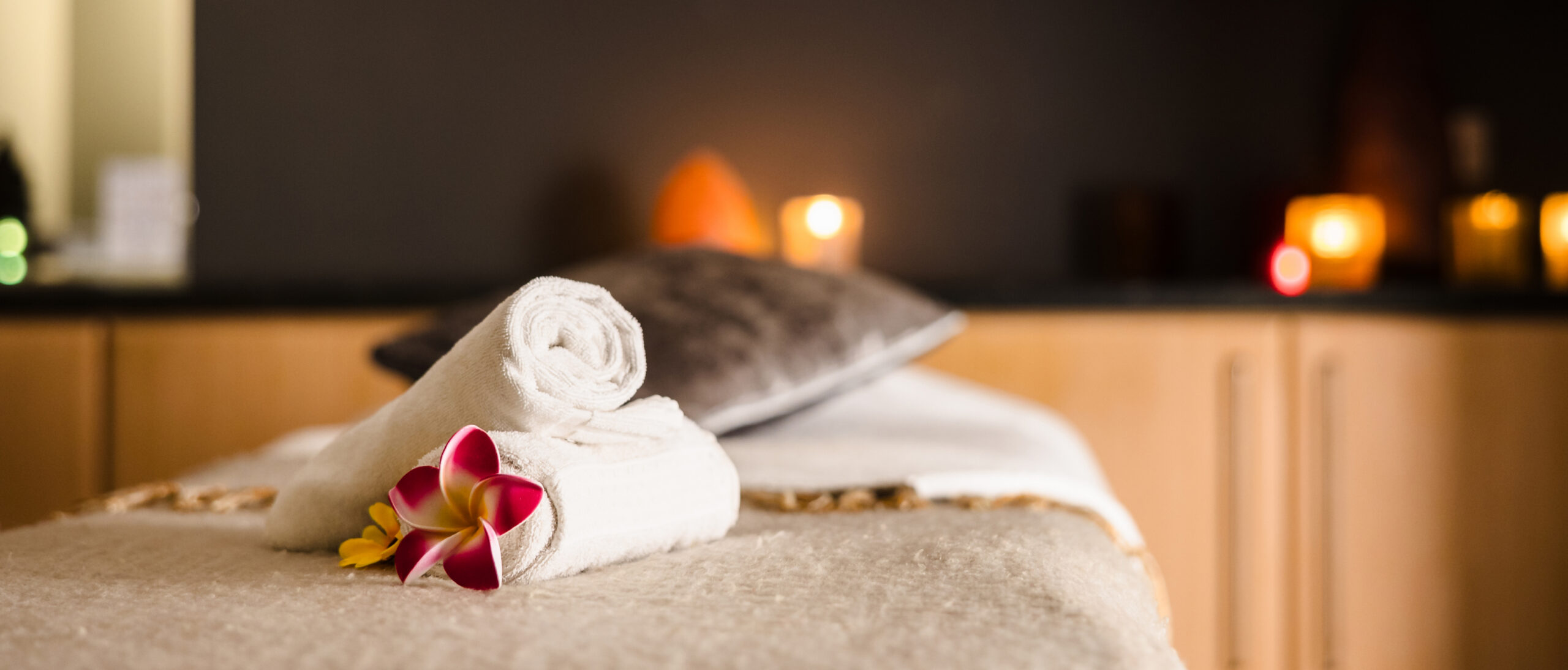 A massage table with rolled white towels and a flower, set in a dimly lit room with candles in the background.