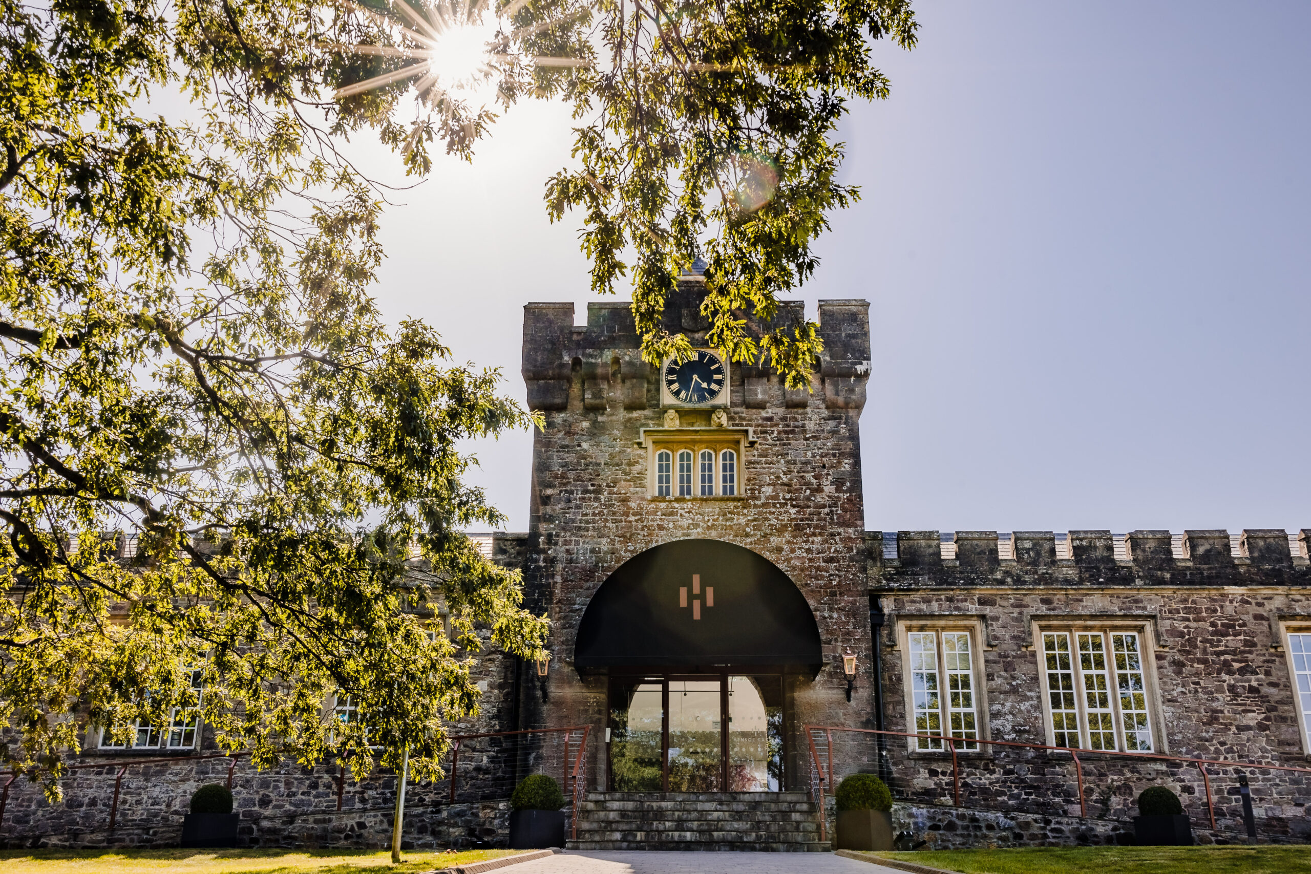 Stone building with a clock tower under a bright sunlit sky, partially framed by tree branches.