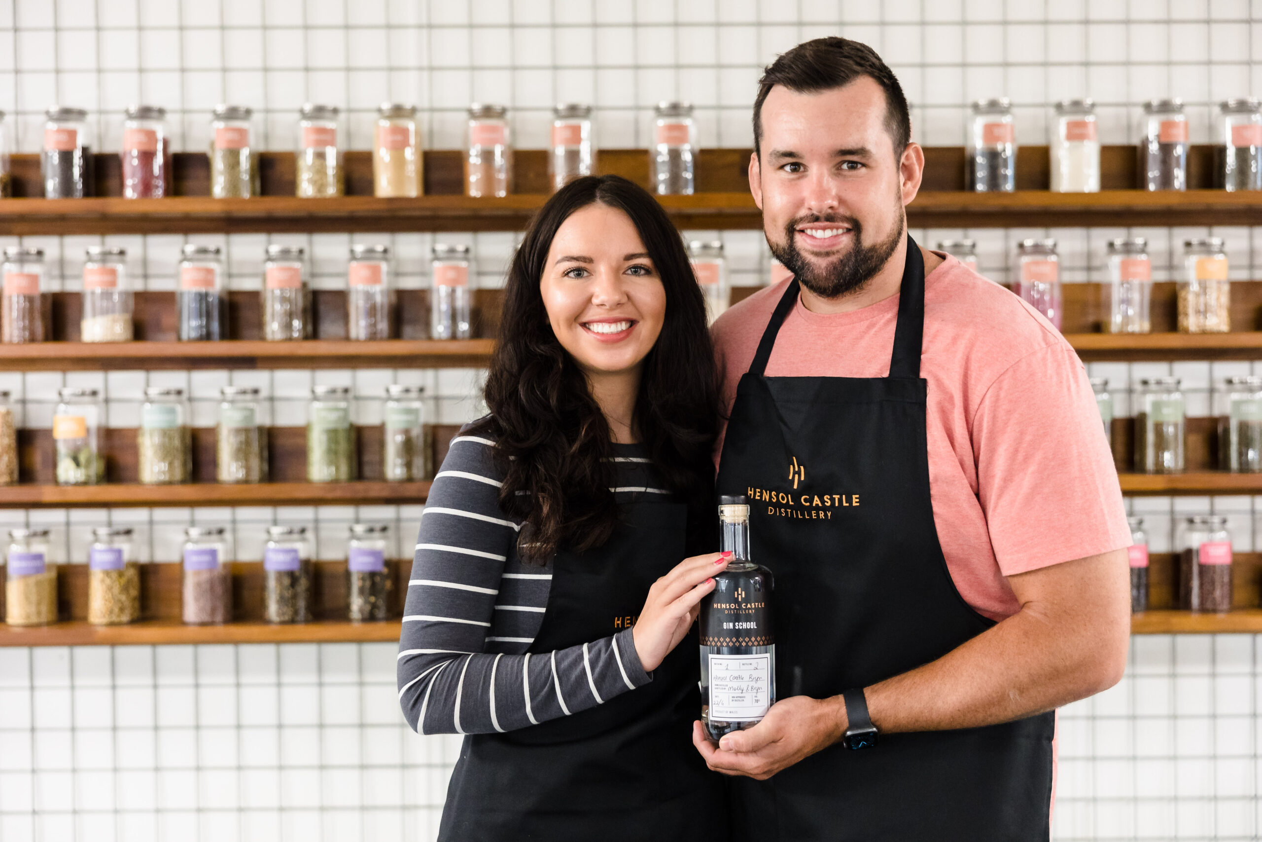 Two people in aprons stand smiling, holding a bottle in front of a wall with shelves of jars.