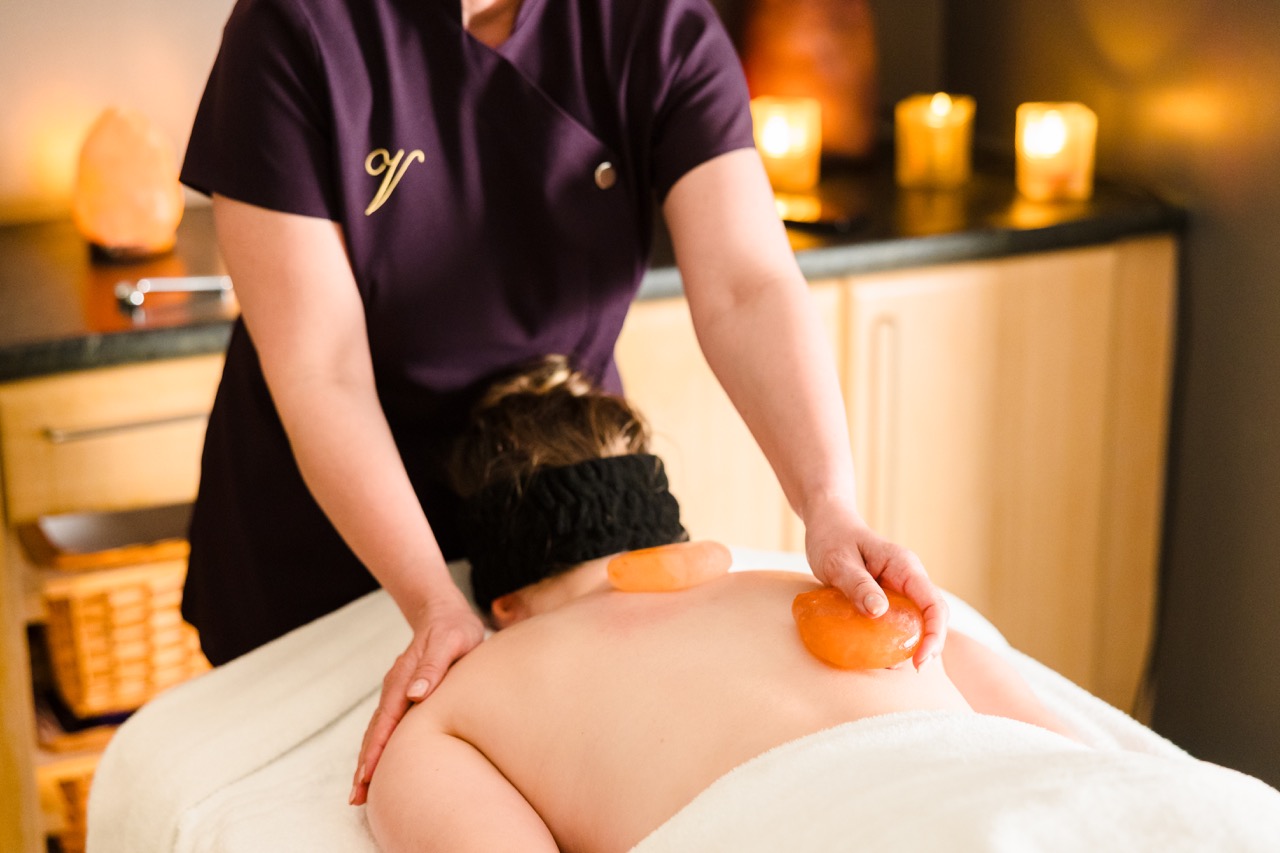 A person receives a hot stone massage in a spa setting, with glowing candles on a nearby counter.