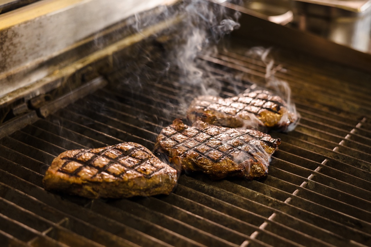 Three pieces of grilled steak with char marks on a grill, emitting smoke.