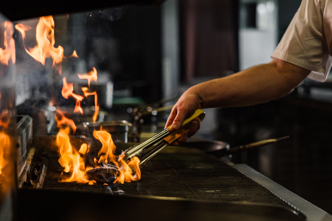 A chef uses tongs to grill meat over open flames in a kitchen.