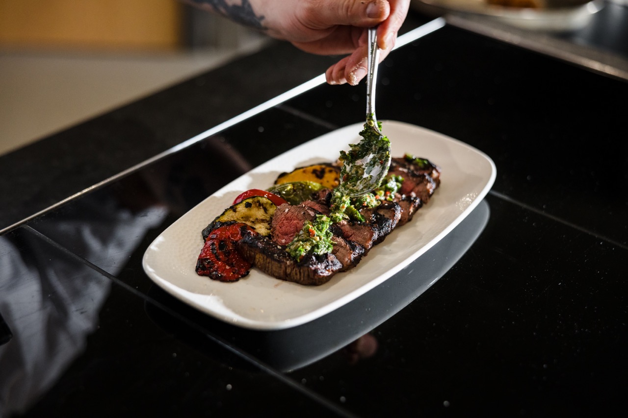 A hand applies green sauce to grilled steak topped with slices of red and yellow roasted vegetables on a white plate.