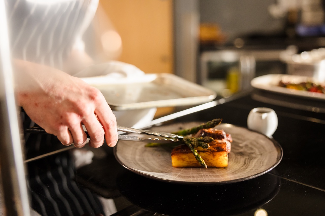 A chef plates a dish with beef, asparagus, and a rectangular piece of food, using tongs on a gray plate in a kitchen setting.