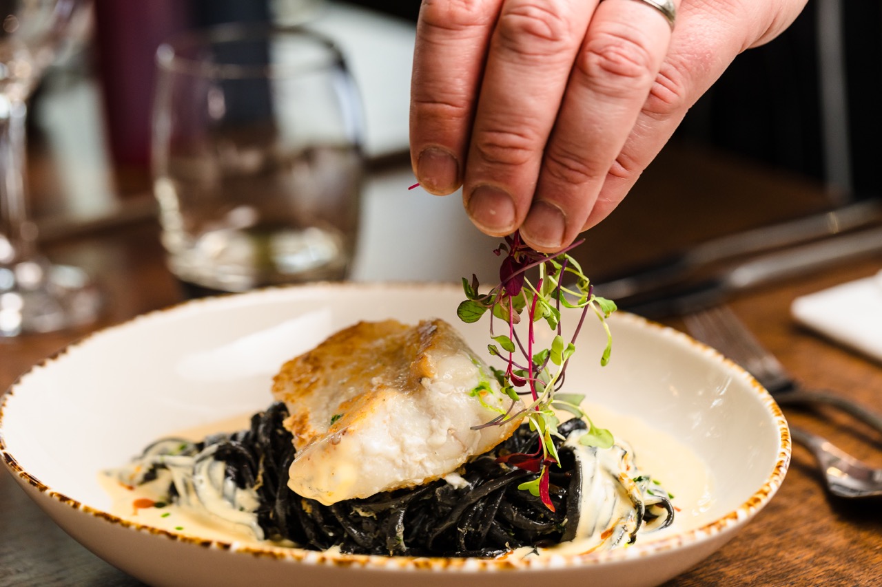 A hand garnishes a plate of squid ink pasta topped with a white fish fillet and sauce.