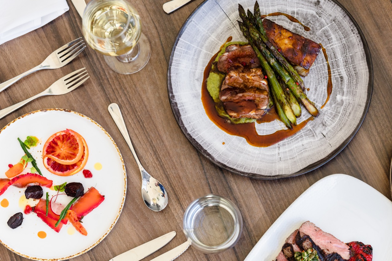 A table with a glass of white wine, a dish of roasted meat and asparagus, a plate of sliced steak, and a salad with blood oranges and berries.