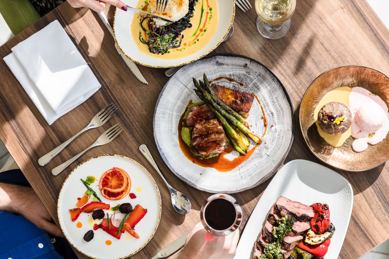 Aerial view of a table with various gourmet dishes, including meat with asparagus, colorful salads, and desserts, accompanied by a glass of red wine and white wine.