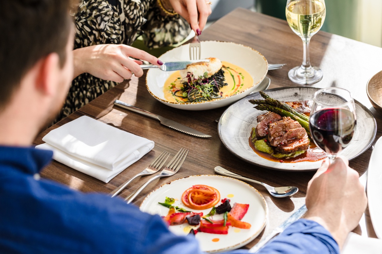 Two people dining at a table with three plated meals, including a meat dish and salad. One person holds a glass of red wine, while the other uses utensils. Glasses of white and red wine are on the table.