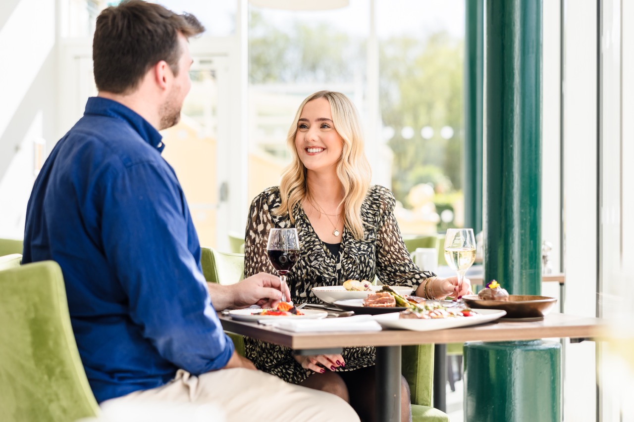 Two people are seated at a restaurant table, enjoying a meal and drinks, with natural light coming through large windows in the background.