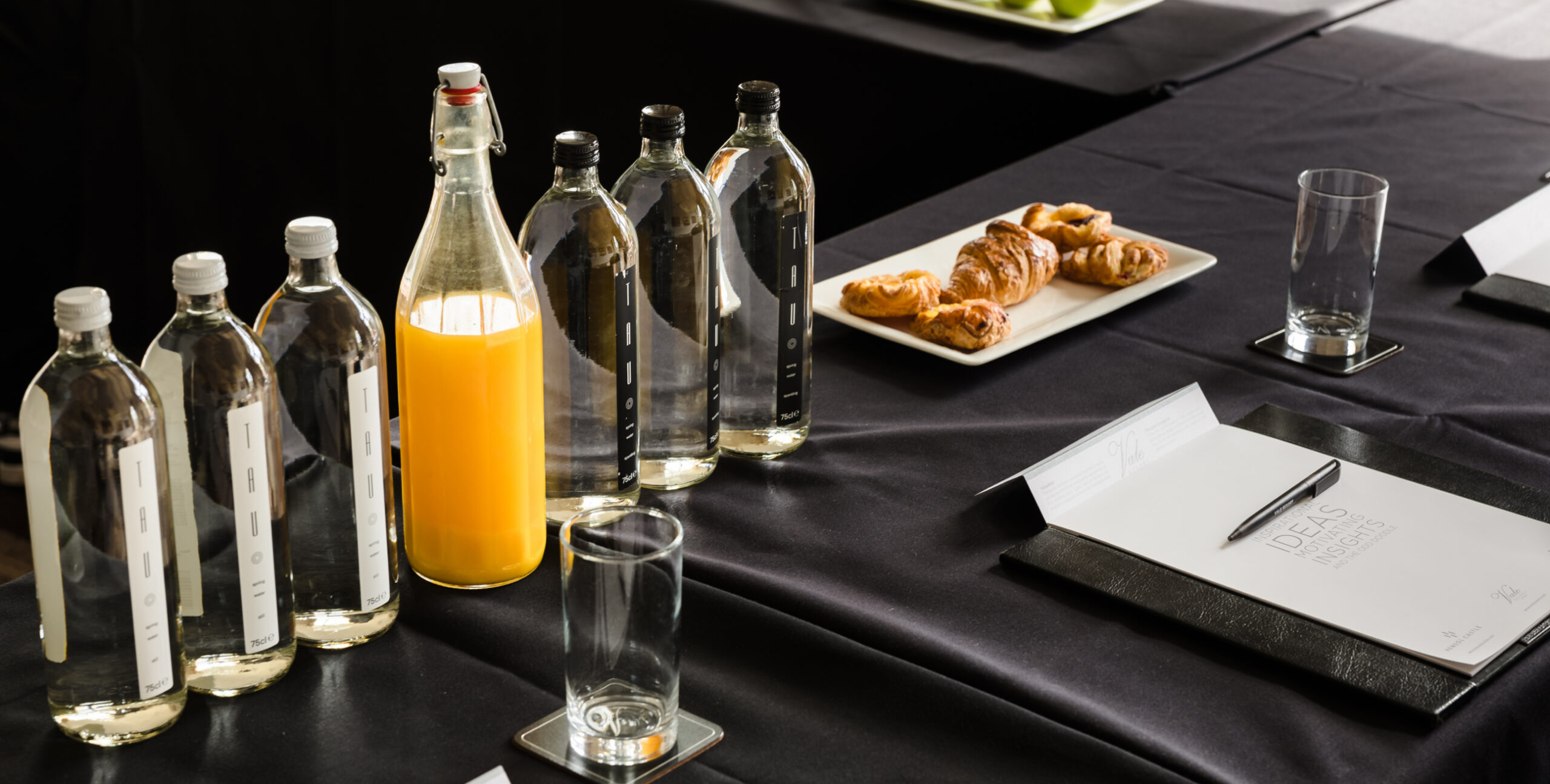 Conference table setup with bottled water, orange juice, croissants, empty glasses, and notepads with pens on a black tablecloth.