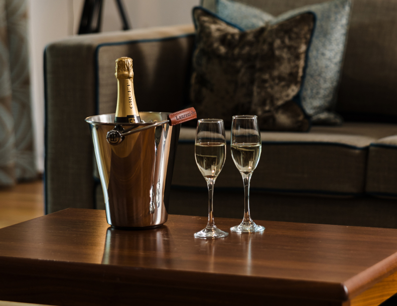 A bottle of champagne in an ice bucket with two filled flutes on a wooden table, next to a gray sofa with cushions.