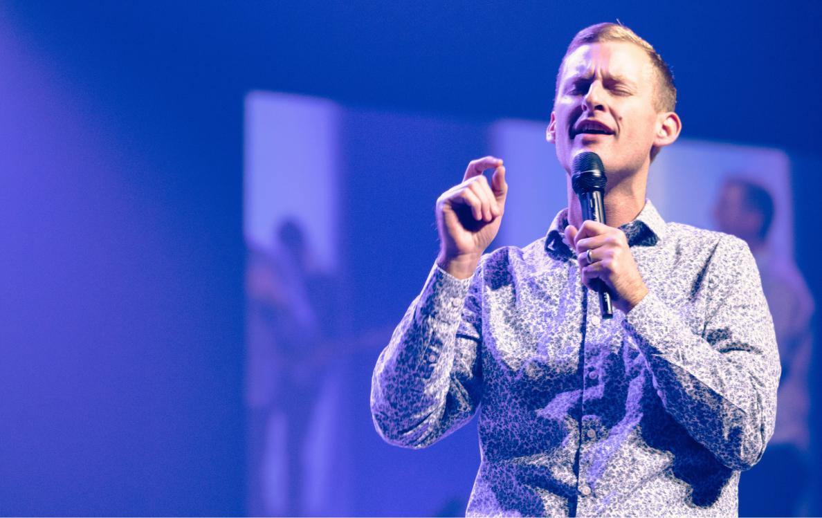 A person is singing passionately on stage, holding a microphone, with a blue background and stage lights.