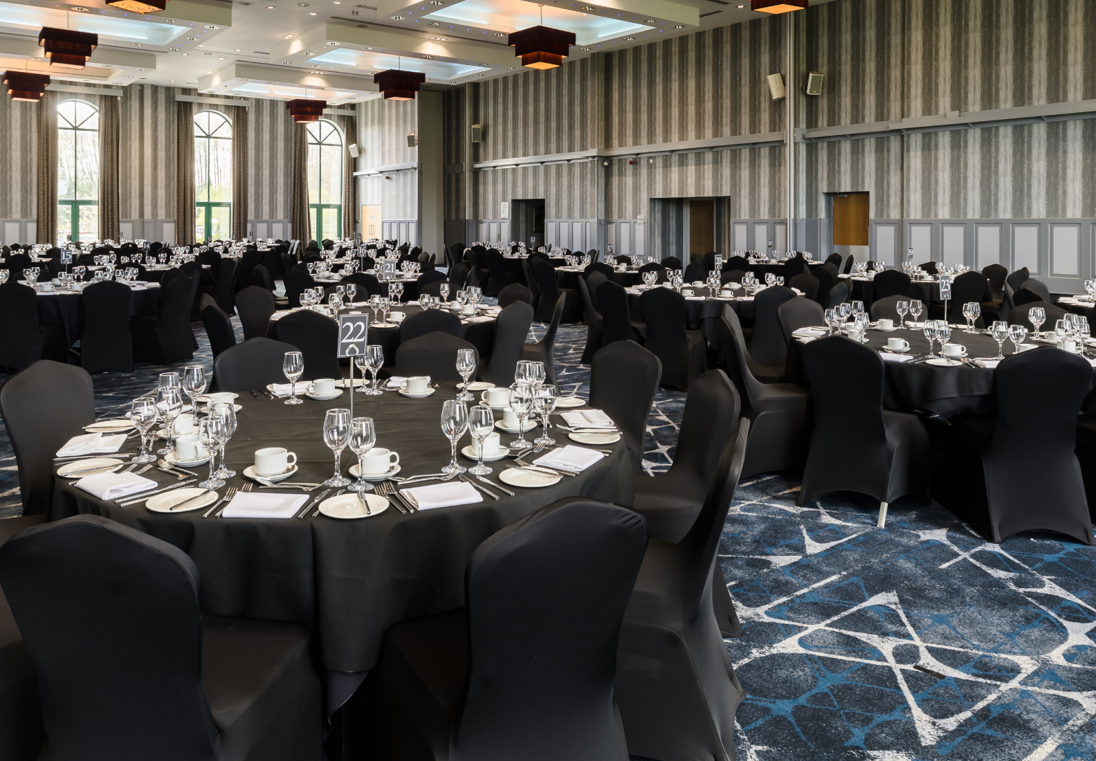 A banquet hall with round tables set with black tablecloths, glassware, and white napkins. Black chair covers and patterned carpet complete the room.