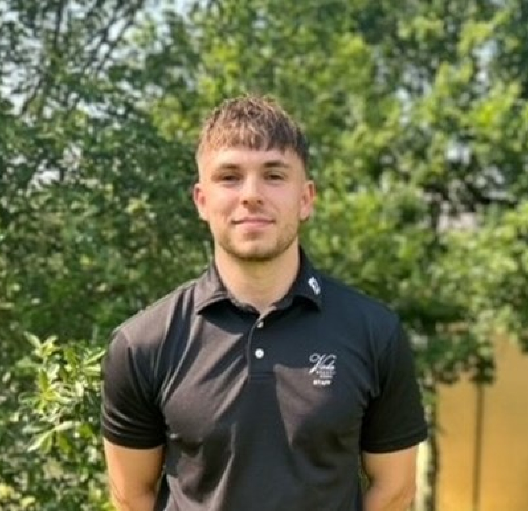 A person stands outdoors wearing a black polo shirt, with greenery in the background.