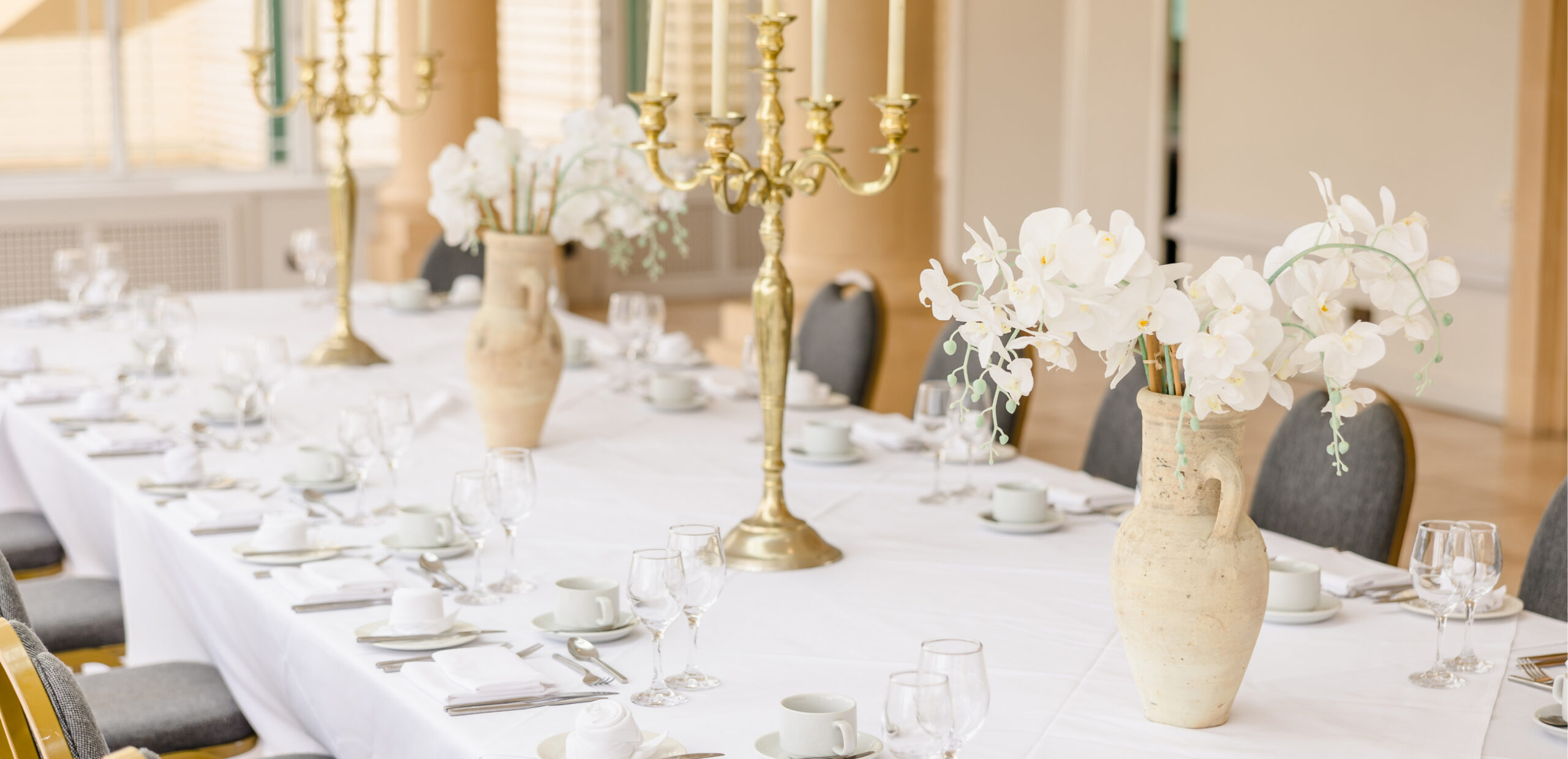 Elegant table setting with white tablecloth, gold candelabras, and vases of white flowers. Dinnerware and glassware are neatly arranged around each seat.