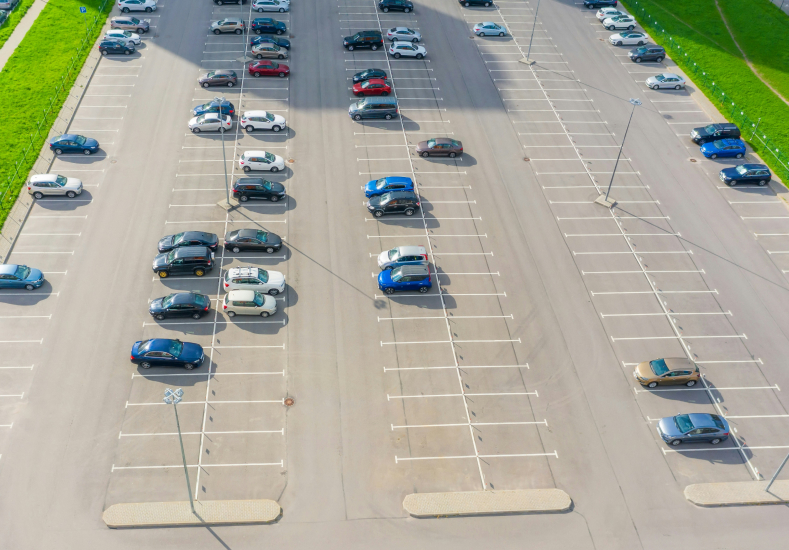 Aerial view of a large parking lot with multiple cars parked in designated spaces. The lot is mostly empty, with several vehicles scattered throughout. Grass borders one side of the area.