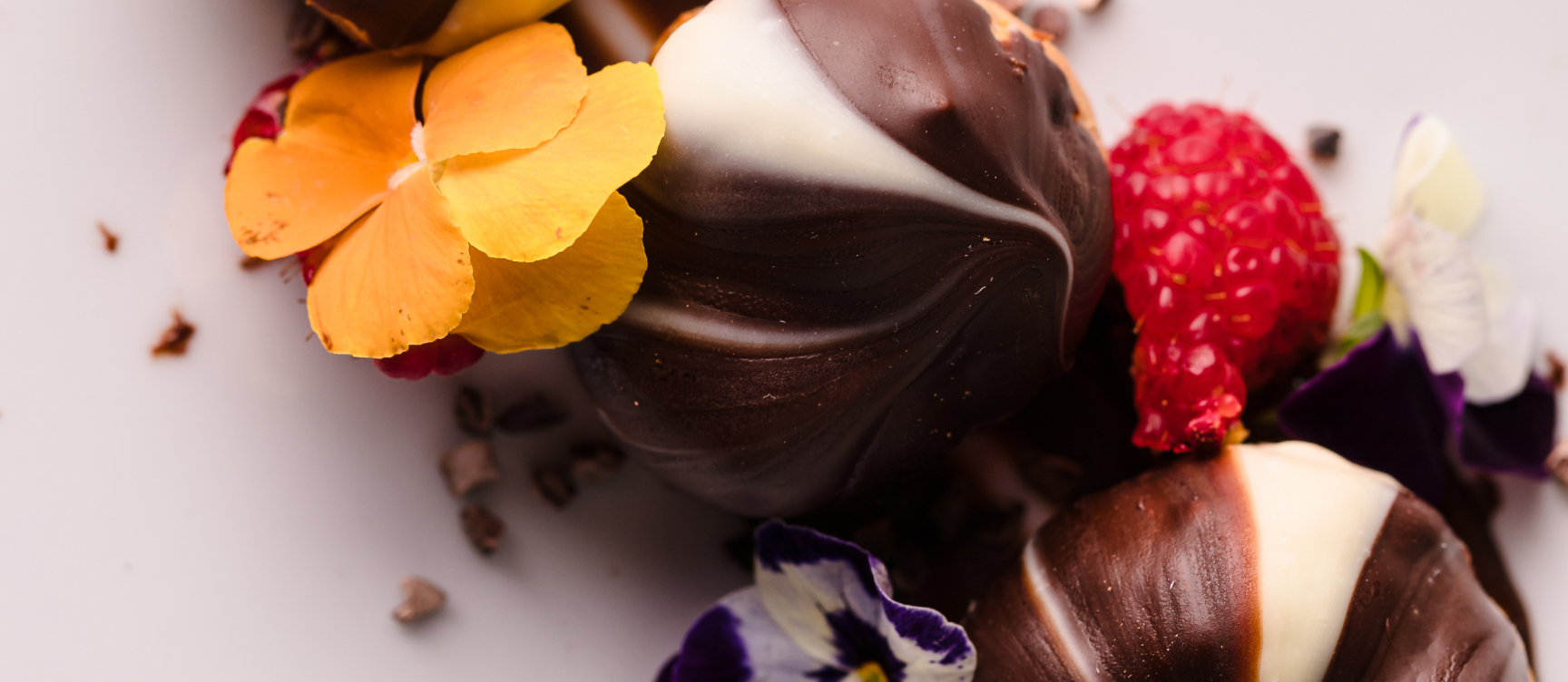 Chocolate truffles with white swirls, garnished with edible flowers, raspberries, and cocoa nibs on a white surface.