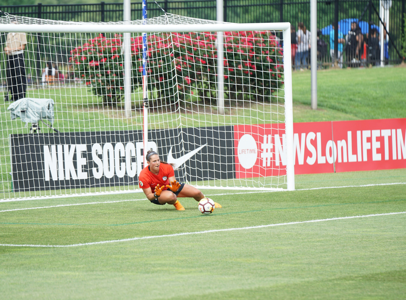 Goalkeeper in an orange jersey crouches low, holding a soccer ball near the goal line on a grass field.