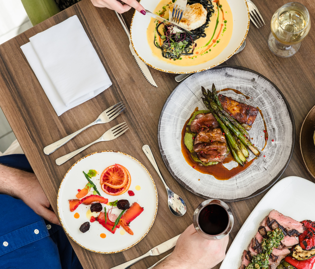 A table with three gourmet dishes, including fish with greens, beef with asparagus, and a colorful citrus salad. A glass of red wine, white wine, and utensils are also visible.