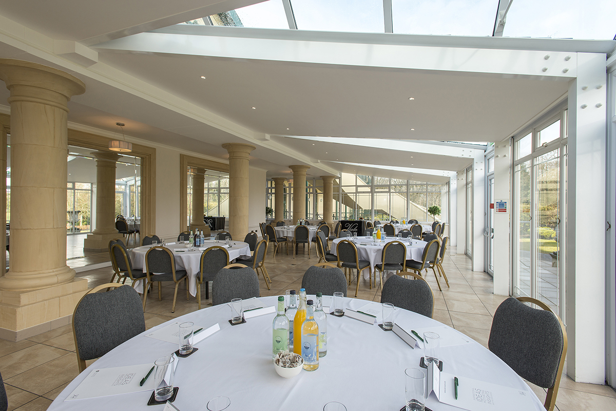 A spacious event room with several round tables covered with white tablecloths, surrounded by chairs. The room has large windows and columns, with bottles and glasses on the tables.