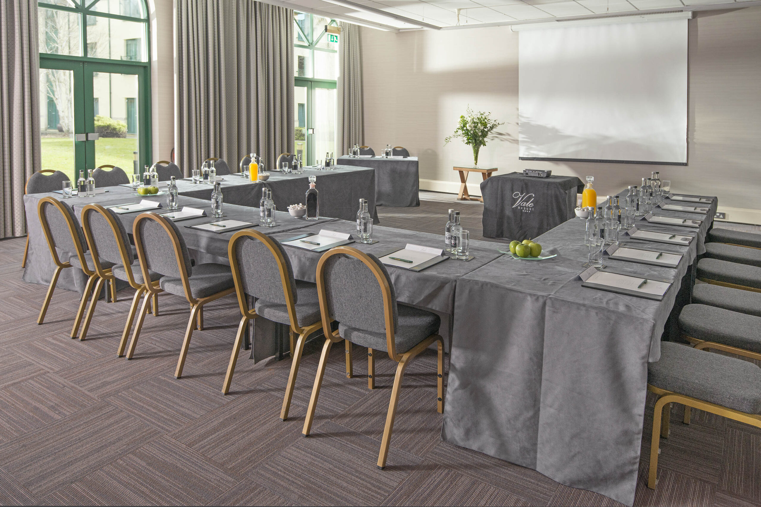 A conference room with U-shaped tables set up, covered in grey cloths, with notepads, pens, and bottles on top. A projector screen and flowers are in the background.