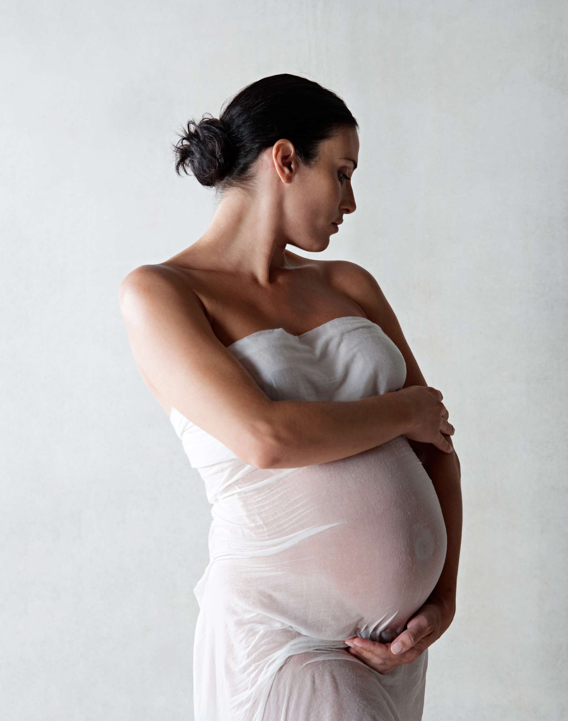 A pregnant person in a white wrap gazes downward while holding their stomach against a plain background.